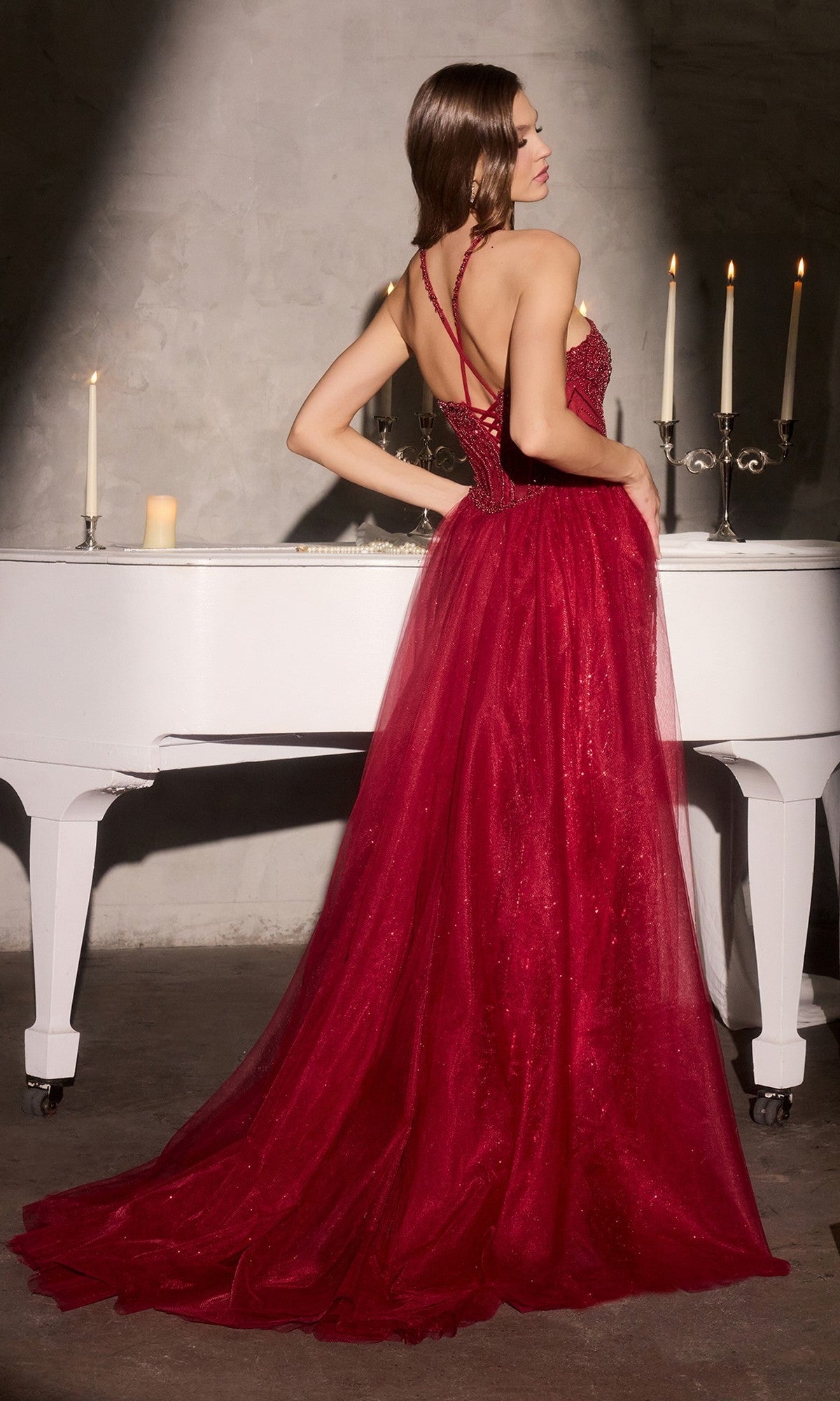 A woman in the Ladivine CC495 long prom dress, featuring a fitted deep red design with a strappy back, stands before a white piano and lit candles against a gray textured wall.